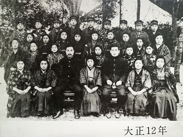 大正12年の記念写真。モノクロ写真で着物姿の生徒たちと着物・学ランを着た先生たちの集合写真