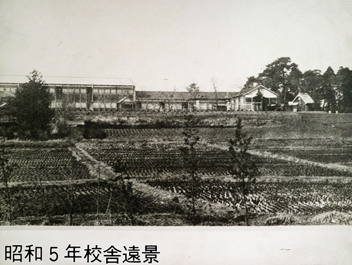 昭和5年校舎遠景。モノクロ写真で手前に田園、その向こうに校舎が写っている